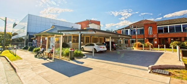 Photo of hospital buildings, parks cars and a woman sitting at a bus stop with a man in a wheelchair